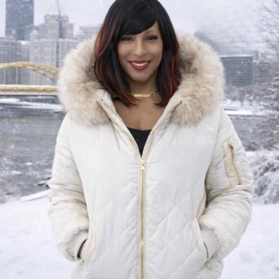 Single in the Steel City 2026 winter portrait of a woman in Pittsburgh wearing a silver bomber jacket with a white fur hood, standing outside during heavy snowfall with a confident smile and red lipstick.